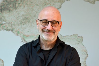 Portrait Peter Korn, schwarzes Hemd, wandfüllende, gräuliche Weltkarte im Hintergrund