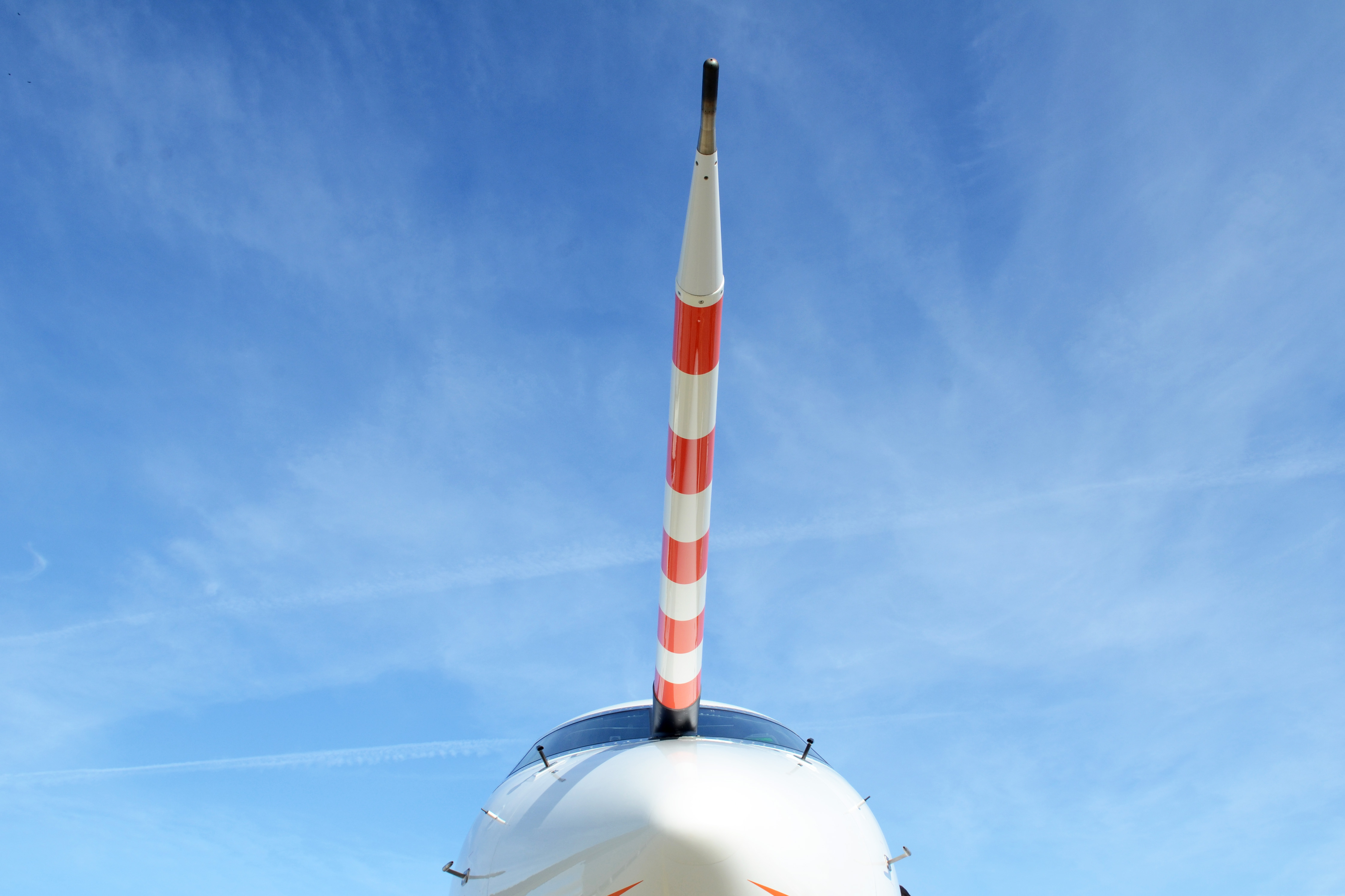 Frontalaufnahme des Flugzeugs HALO, dominiert durch den rot-weiß gestreiften Nasenmast vor blauem Himmel mit leichter Bewölkung.