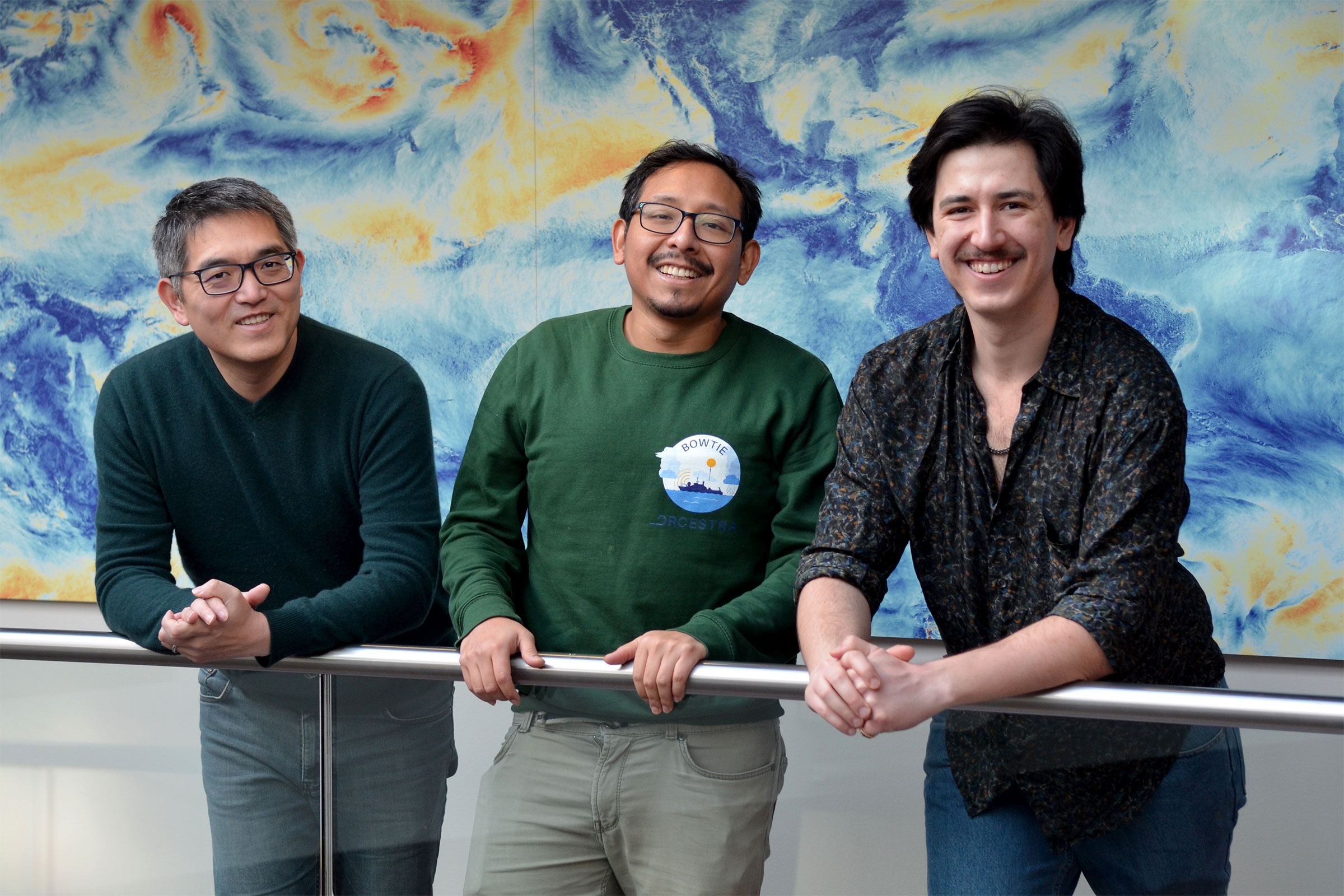 Chao Li, Hans Segura und Noel Brizuela am Max-Planck-Institut für Meteorologie. Sie stehen an einem Geländer vor einer großformatigen und ästhetischen Visualisierung.