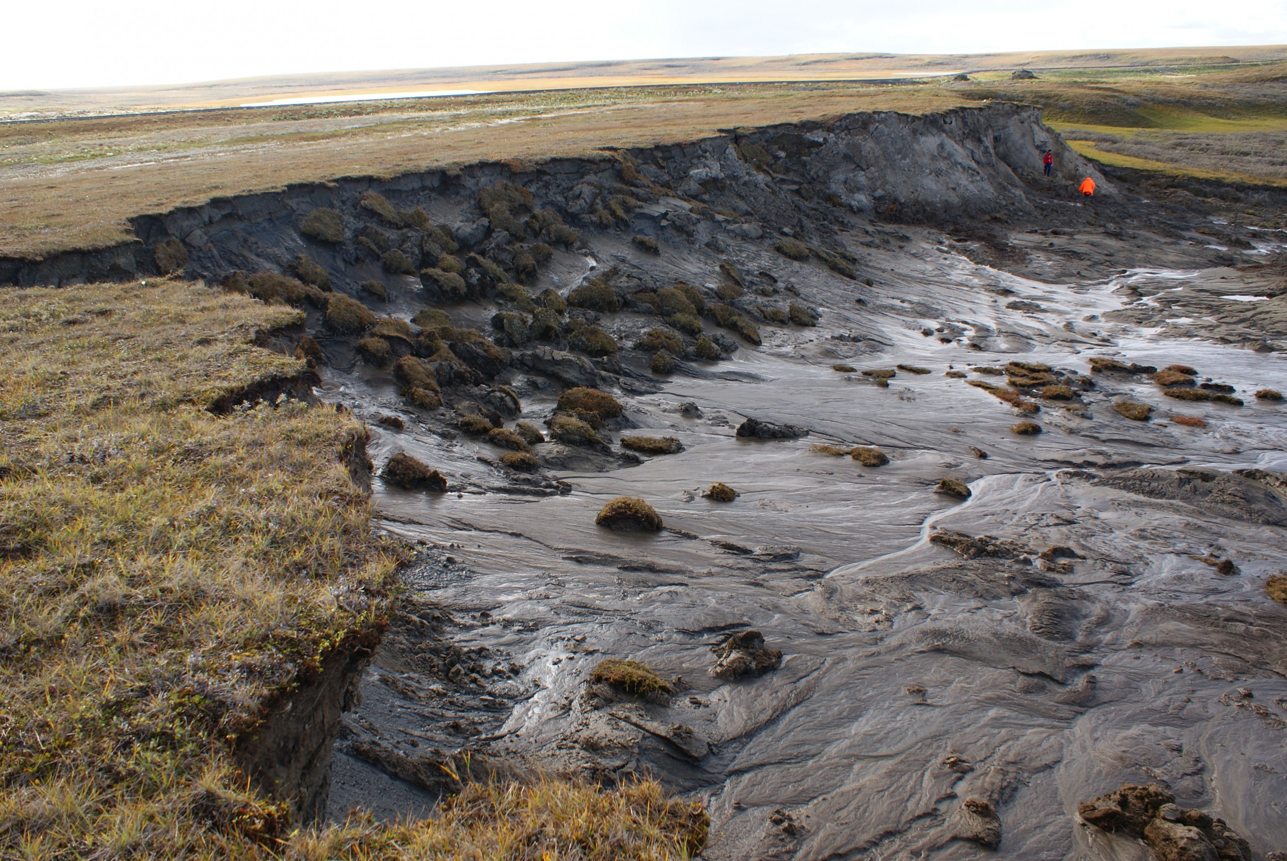 Photograph of a thawing permafrost area in Siberia. The landscape shows a steep erosion edge with slumped soil and peat blocks, as well as flowing mud patterns downhill. In the background, two people in bright clothing are examining the site. The surrounding area is flat and tundra-like, with sparse vegetation.