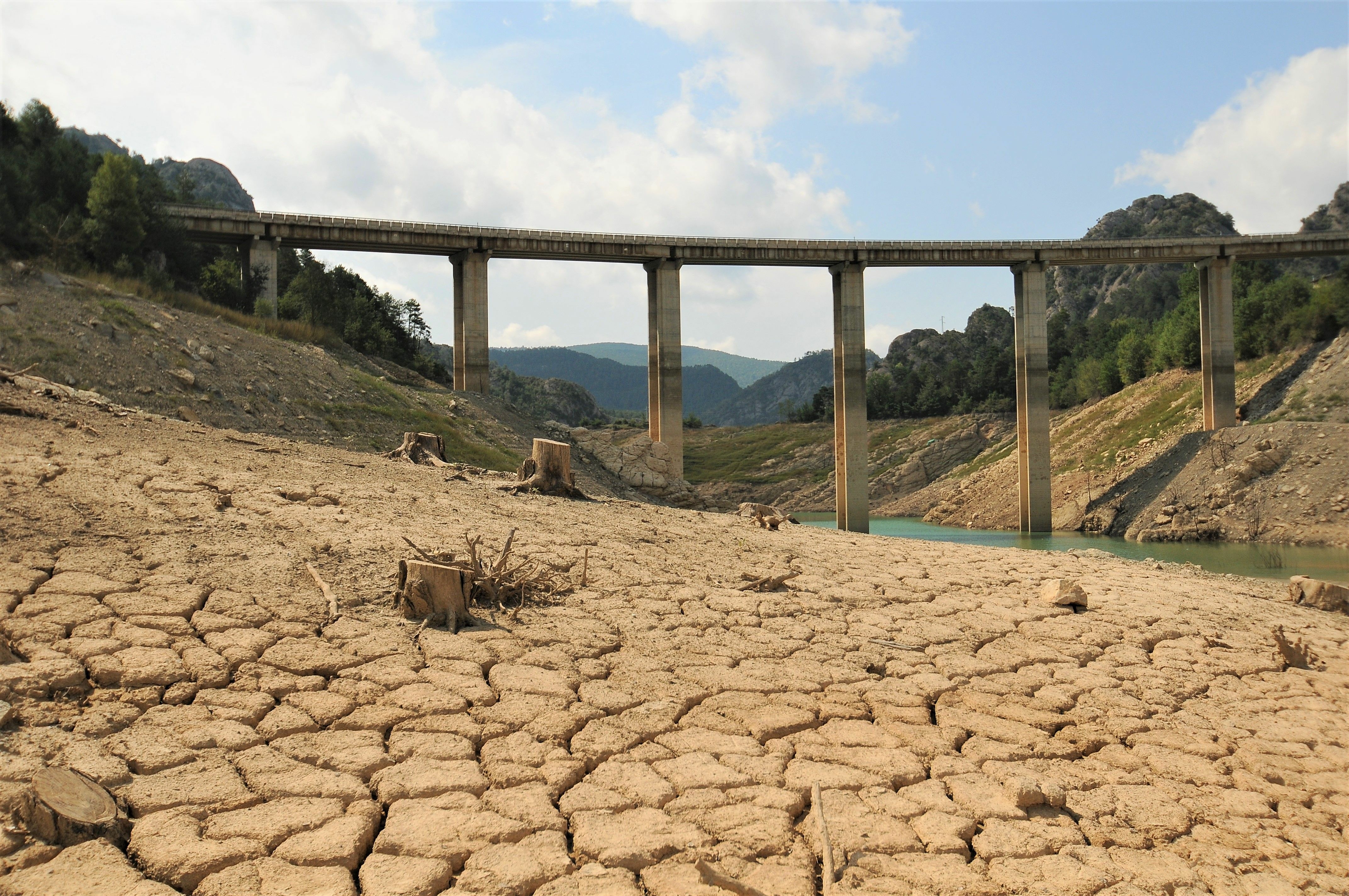 A dry landscape with cracked soils, a bridge leads across a river with very low water level.