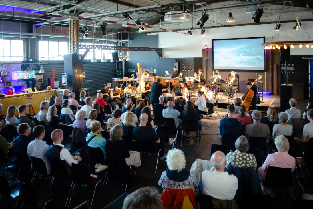 Blick durch einen Veranstaltungsraum über das Publikum von hinten zur Bühne auf der ein Orchester spielt. Im Hintegrund, an der Wand hinter dem Orchester hängt ein großer Bildschirm auf dem 2 große Wellen auf einem Meer zu sehen sind.