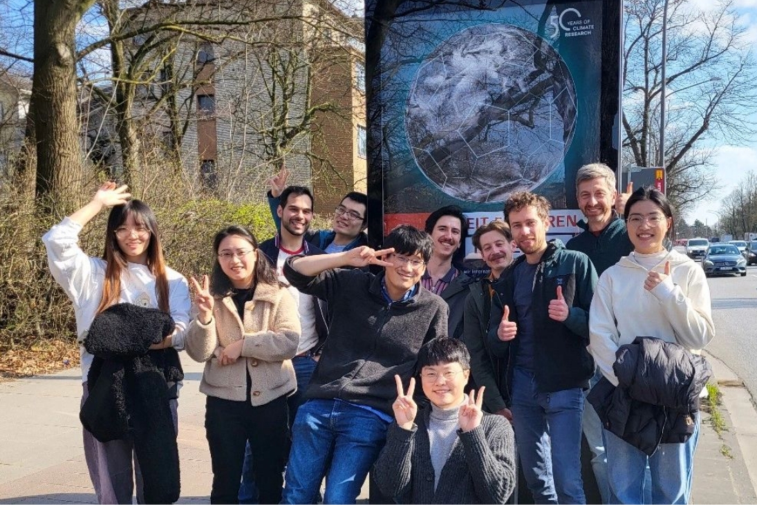 Gruppenfoto von 11 Personen, die auf einer Straße vor einer Plakatwand mit dem Plakat zum Jubiläum des MPI-Ms stehen. Sie lächeln und winken.