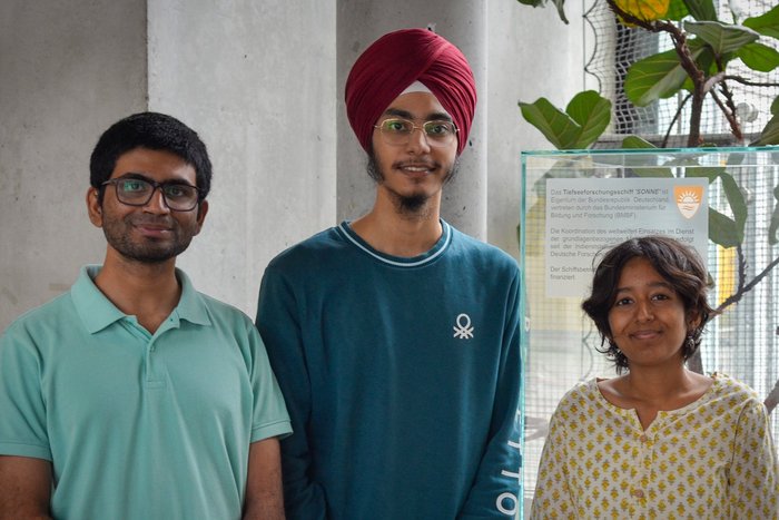 group of 3 people in front of a predominantly gray background, 2 people on the left with glasses, the person on the right is the smallest, person in the middle wears a turban