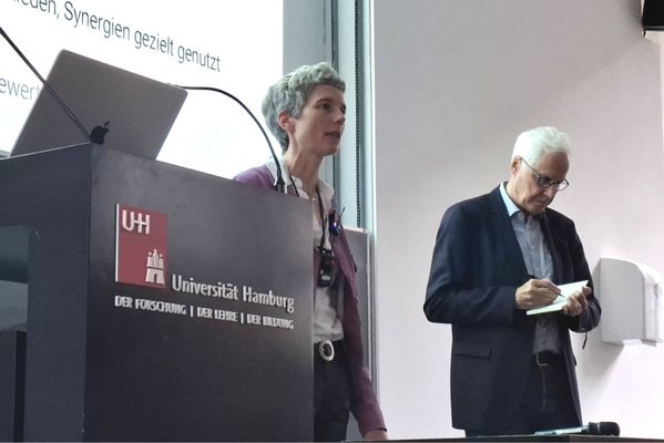 2 Personen stehen vor einer Wand auf die eine Päsentation projiziert ist. Links ist ein Stehpult mit dem Logo und dem Schriftzug der Universität Hamburg zu sehen. Dei Person rechts trägt eine Brille und schreibt in ein aufgeschlagenes Buch.
