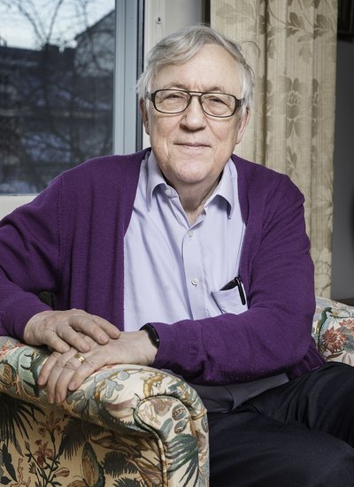 Lennart Bengtsson sitzt in einem Polstersessel vor einem geschlossenen Fenster und einem goldenen Vorhang. Er trägt eine lila Strickjacke. Er blickt direkt in die Kamera.