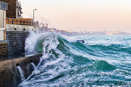 Türkisgrüne Brandungswelle schlägt an eine steinerne Ufermauer mit Abflussöffnungen; Wasser läuft über die Promenade, links Häuser, rechts offenes Meer, am Horizont eine Küstenstadt unter rosa‑blauem Himmel.