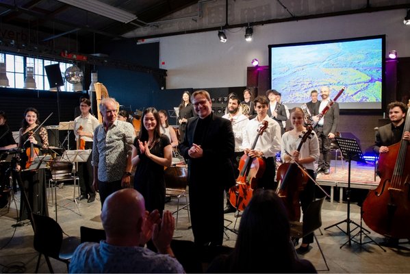 Ein stehendes Orchester und 3 Personen im vor dem Orchester empfangen Applaus eines Publikums von dem 2 Personen von hinten im Vrodergrund zu sehen sind. Im Hintergrund der ühne an der Wand hängt ein großer Bildschirm auf dem eine grüne Landschaft aus der Vogelperspektive gezeigt wird.
