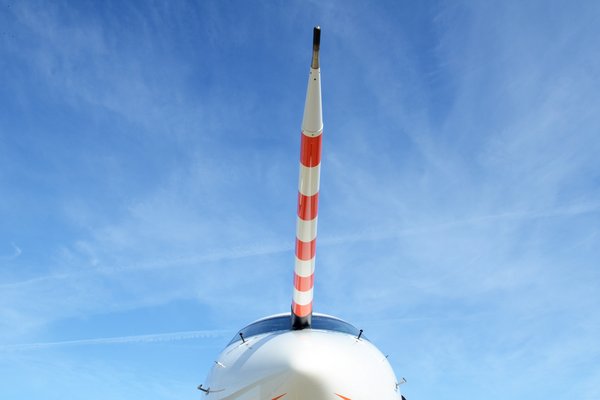 Frontalaufnahme des Flugzeugs HALO, dominiert durch den rot-weiß gestreiften Nasenmast vor blauem Himmel mit leichter Bewölkung.