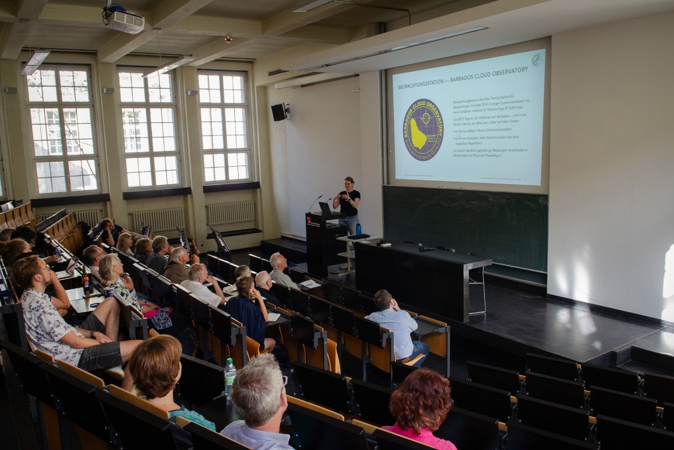 Vorlesung in einem Hörsaal. Raum steigt zum Kamerastandort hin an. Zuhörer sind von hinten zu sehen. Die vortragende Person ist im Zentrum des Bilds kaum ausnehmbar. Eine helle Vortragsfolie wird im Hintergrund an die Wand projiziert.