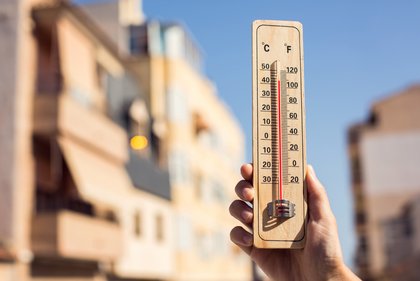 Eine Hand hält ein Holzthermometer mit rotem Quecksilberstrang, das Temperaturen in Grad Celsius und Fahrenheit anzeigt. Der rote Strang zeigt etwa 40 °C (100 °F) an. Im unscharfen Hintergrund ist ein Gebäude unter blauem Himmel zu sehen, was auf eine heiße Außentemperatur hindeutet.