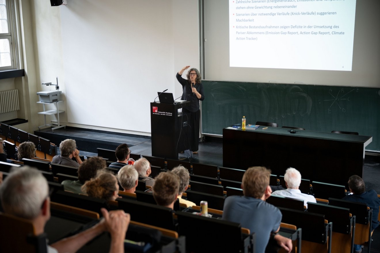 Vorlesung in einem Hörsaal. Raum steigt zum Kamerastandort hin an. Zuhörer sind von hinten zu sehen. Die vortragende Person steht hinter einem schwarzen Stehpult, blickt ins Publikum, trägt Brille, hält mit der linken Hand ein Mikrofon, hebt den rechten Arm in Kopfhöhe.